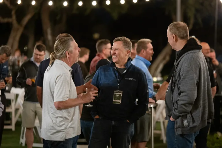 Group of middle-aged men socializing outdoors at a nighttime event with string lights overhead, some holding drinks and wearing casual jackets and shirts.