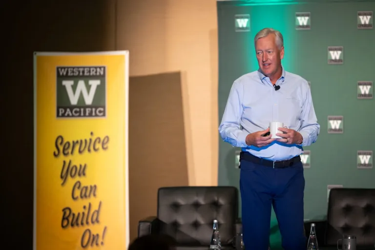 A man in a light blue shirt and dark blue pants holding a white mug and speaking on a stage with Western Pacific logos on green backdrop and a yellow banner that reads "Service You Can Build On!".