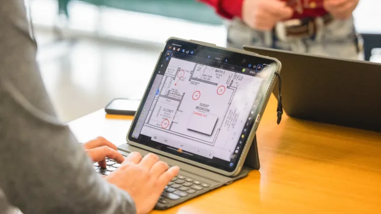 Person typing on a keyboard case attached to a tablet displaying a digital floor plan marked with red annotations on a wooden table, with another person and a closed laptop in the background.