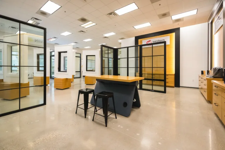 Modern showroom interior featuring large glass window displays with black frames, a central wooden table with two black metal stools, light wooden cabinetry, and bright ceiling lights.