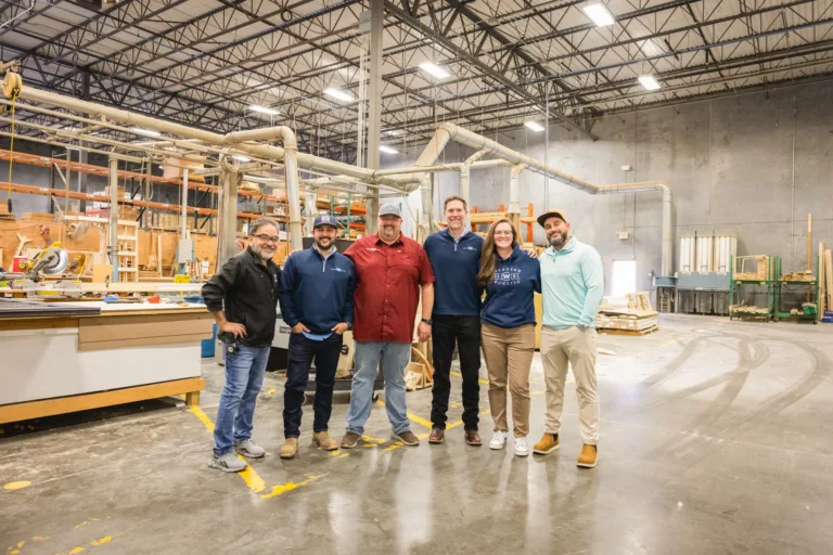 Six people standing in a large industrial workshop with machinery and wooden materials in the background, posing for a group photo.