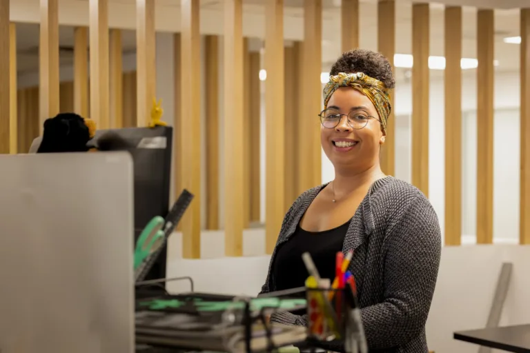 Smiling woman with glasses and a patterned headband sitting at a desk in a modern office with wooden vertical slats in the background.