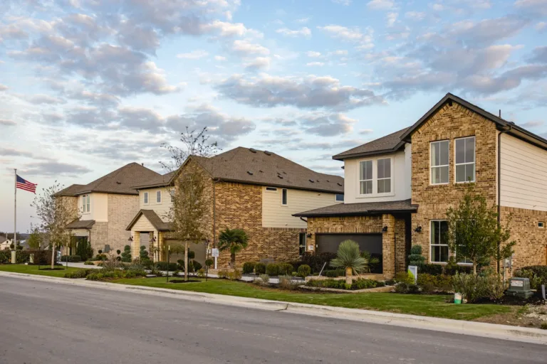 Row of modern suburban two-story houses with brick and stone facades, neatly landscaped yards, and an American flag on a pole under a partly cloudy sky.