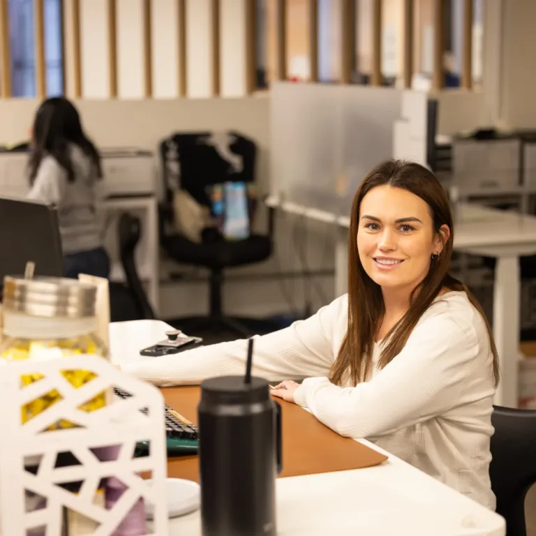 Smiling woman with long brown hair sitting at a desk in an office environment with computer monitors and office supplies around her.